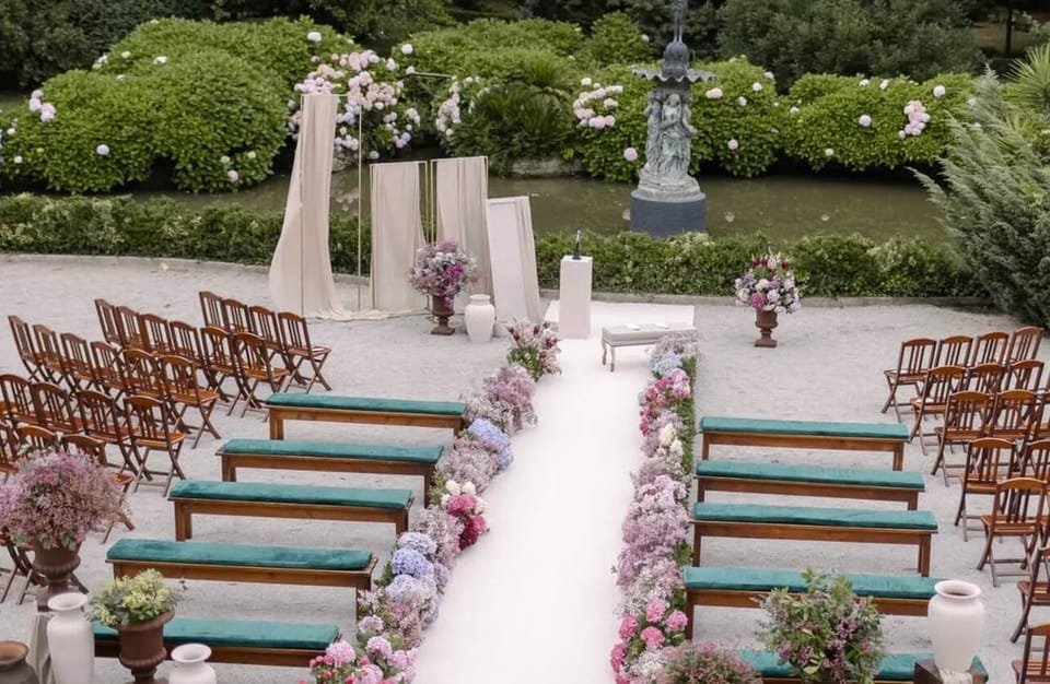 Cerimónia num jardim: corredor branco ladeado por flores em tons pastel, bancos com almofadas verdes, cadeiras de madeira e painéis de tecido claro junto ao altar.