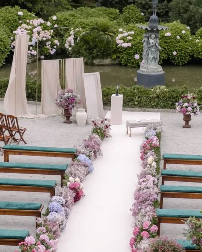 Cerimónia num jardim: corredor branco ladeado por flores em tons pastel, bancos com almofadas verdes, cadeiras de madeira e painéis de tecido claro junto ao altar.