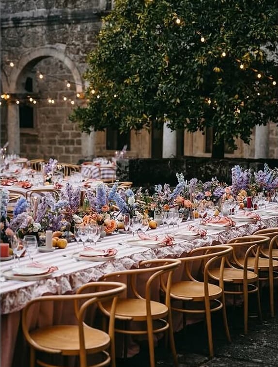 Díptico: à esquerda, mesa comprida com flores em lilás, azul e laranja e cadeiras de madeira curvada; à direita, mesas redondas com toalhas às riscas num claustro de pedra iluminado por cordões de lâmpadas.