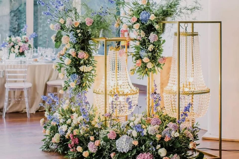 Instalação floral em rosa, azul e branco sobre estruturas douradas com lustres de cristal, numa sala luminosa com chão de madeira.