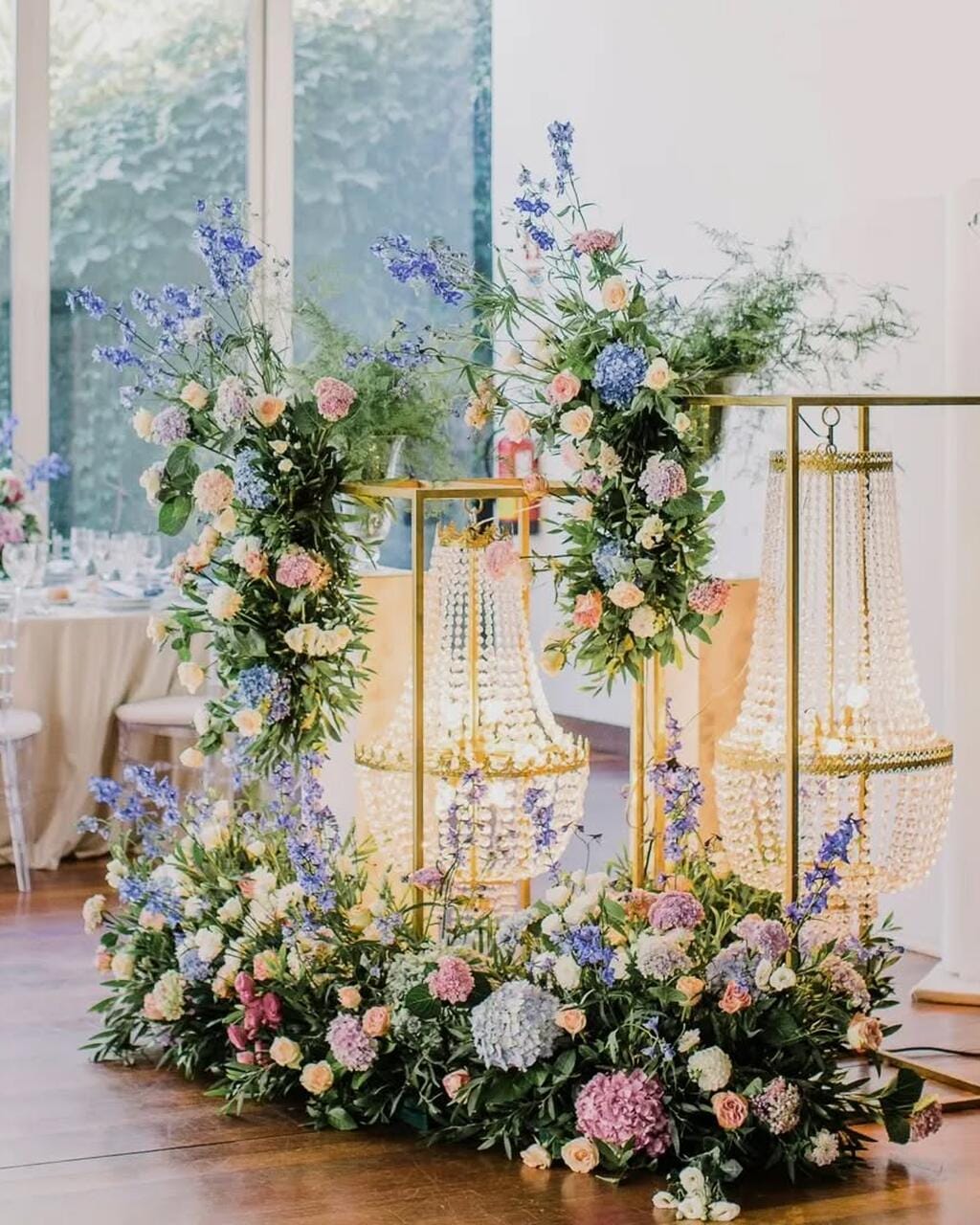 Instalação floral em rosa, azul e branco sobre estruturas douradas com lustres de cristal, numa sala luminosa com chão de madeira.