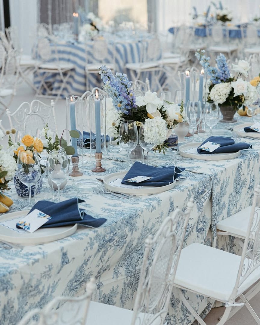 Mesa posta com toalha estampada em azul e branco, guardanapos azul-escuros, velas azuis e arranjos de flores brancas, azuis e amarelas com limões.