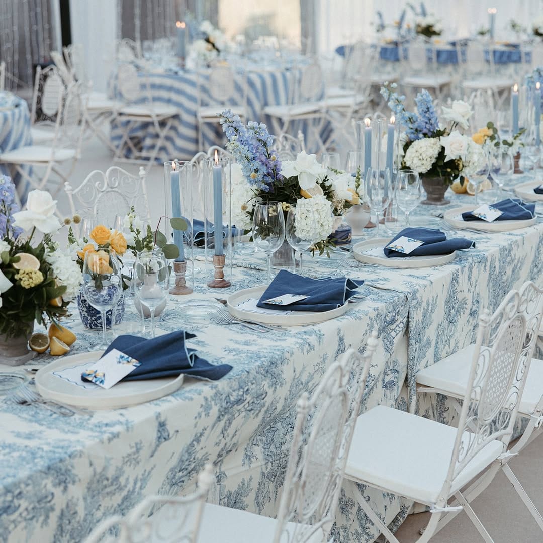 Mesa posta com toalha estampada em azul e branco, guardanapos azul-escuros, velas azuis e arranjos de flores brancas, azuis e amarelas com limões.