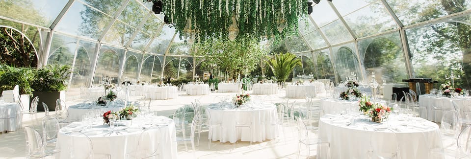 Sala de receção numa estrutura transparente, com mesas redondas de toalha branca, cadeiras transparentes, centros de flores vermelhas e rosa e uma instalação de verde suspensa com lustres.