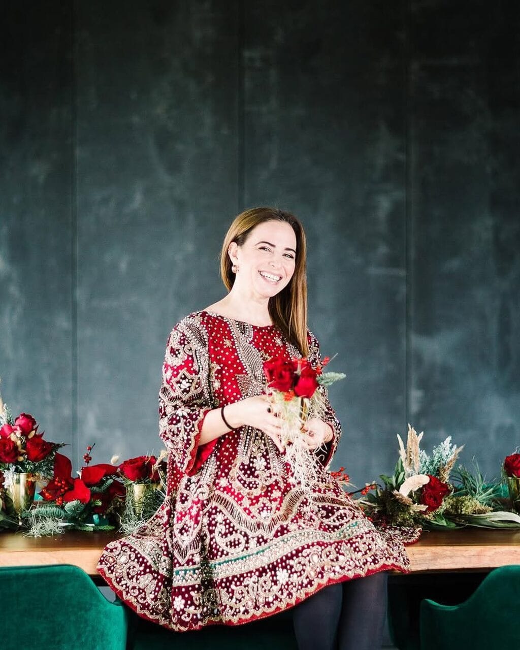 Retrato de uma mulher sorridente, sentada numa mesa de madeira entre arranjos de rosas vermelhas e verdes, diante de uma parede verde-escura.